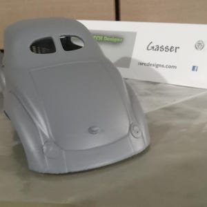 Pu&ograve; includere: Una carrozzeria per modellino d'auto grigia non verniciata con cofano arrotondato, due rientranze circolari per i fari e un abitacolo con due piccole aperture per i finestrini. Un cartoncino bianco riporta il testo Gasser e ssrcdesigns.com.
