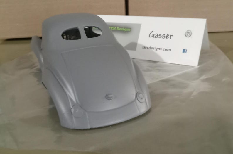Pu&ograve; includere: Una carrozzeria per modellino d'auto grigia non verniciata con cofano arrotondato, due rientranze circolari per i fari e un abitacolo con due piccole aperture per i finestrini. Un cartoncino bianco riporta il testo Gasser e ssrcdesigns.com.