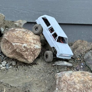 Könnte beinhalten: Ein hellblaues Spielzeugauto mit großen, strukturierten Reifen ist auf einem felsigen Gelände abgewinkelt. Der Truck ist so positioniert, als würde er über einen großen Felsen klettern. Die Szene spielt sich im Freien ab, mit einem grauen Hintergrund.