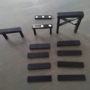 May include: A collection of small, black wooden furniture pieces, including tables and supports. The set includes a small table with two legs, a table with four legs, and a sawhorse-style support. Several rectangular pieces are also present.