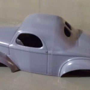 Pu&ograve; includere: Una carrozzeria per modellino d'auto di colore grigio con tetto arrotondato, parafanghi curvi e aperture per i finestrini. Questo componente in plastica non verniciata mostra la silhouette classica di una coup&eacute; d'epoca con cofano e bagagliaio.