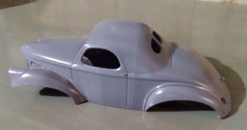 Pu&ograve; includere: Una carrozzeria per modellino d'auto di colore grigio con tetto arrotondato, parafanghi curvi e aperture per i finestrini. Questo componente in plastica non verniciata mostra la silhouette classica di una coup&eacute; d'epoca con cofano e bagagliaio.