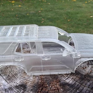 May include: A translucent, unpainted model car body shell featuring a detailed SUV silhouette with a roof rack, side windows, and wheel arches. The clear, hollow plastic structure displays the exterior contours of a vehicle, including the hood, roof, and side panels, designed for assembly or customization.