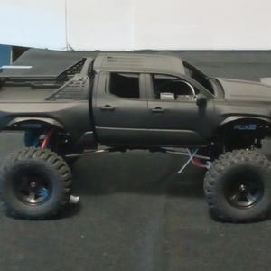 Può includere: Un modello di pick-up radiocomandato nero opaco con grandi pneumatici fuoristrada, un sistema di sospensioni rialzato con componenti blu e rossi visibili e la scritta FCX18 stampata in blu sull'area del passaruota anteriore del veicolo.