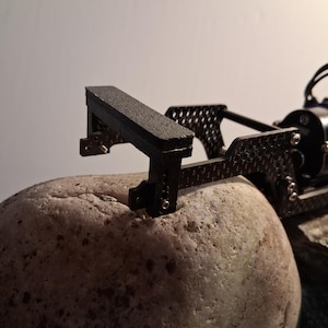 May include: A black carbon fiber chassis component for a remote-controlled vehicle, featuring a textured rectangular bumper, metallic screws, and a blue electric motor attached to the rear frame, resting on a large, smooth, light-colored stone.