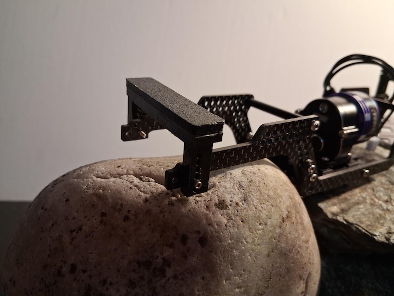 May include: A black carbon fiber chassis component for a remote-controlled vehicle, featuring a textured rectangular bumper, metallic screws, and a blue electric motor attached to the rear frame, resting on a large, smooth, light-colored stone.