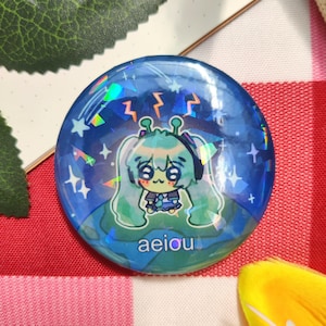 Op de afbeelding: Een ronde, blauwe button met een holografisch effect. Het toont een cartoon alien karakter met groen haar, een koptelefoon en een blauwe outfit. De button heeft het woord "aeiou" onderaan en is versierd met sterren en bliksemschichten.