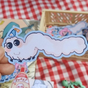 May include: A die-cut sticker featuring a pale pink, glittery, worm-like creature with a blue outline. The creature has large dark eyes, a small smiling mouth, and wears a tiny blue party hat with orange accents. It has several small black legs.