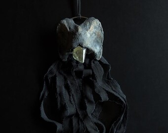 May include: A black fabric sculpture with a gray, textured, bird-like head and a clear crystal beak. The fabric is frayed and hangs in long strands.