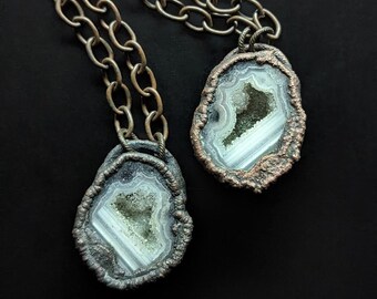 May include: Two silver chain necklaces with agate pendant charms. The agate is a light grey with a white and green crystal inside. The pendants are set in a copper-toned metal.