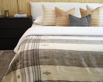 May include: A bed with a white headboard and a patterned blanket in shades of gray and cream. Several pillows in neutral tones are arranged on the bed. A black nightstand with a lamp and books sits beside the bed. The wall behind the bed is made of bamboo.