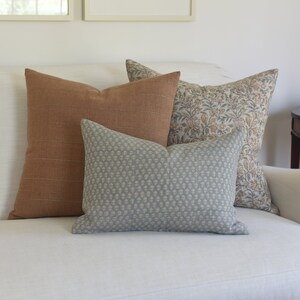 May include: Three decorative throw pillows on a white sofa. The pillows are a rust orange, a light blue with a white pattern, and a floral pattern in shades of brown, beige, and white.