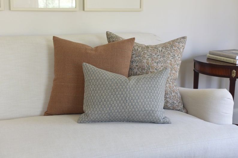 May include: Three decorative throw pillows on a white sofa. The pillows are a rust orange, a light blue with a white pattern, and a floral pattern in shades of brown, beige, and white.