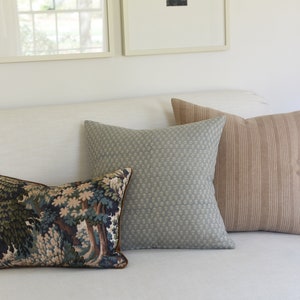 May include: Three decorative throw pillows on a white couch. The pillows are a tapestry pattern with trees, a light blue patterned fabric, and a brown and white striped fabric.