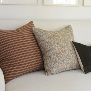 May include: Three decorative throw pillows on a white sofa. The pillows are brown with white stripes, beige with a floral pattern, and dark green with gold accents.