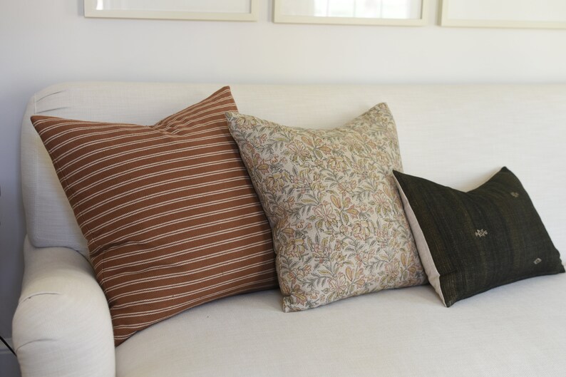 May include: Three decorative throw pillows on a white sofa. The pillows are brown with white stripes, beige with a floral pattern, and dark green with gold accents.