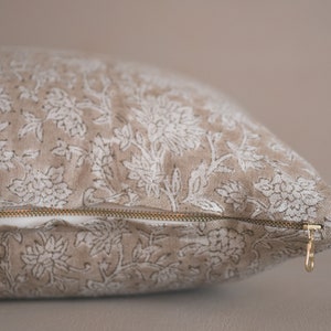 Puede incluir: Una almohada beige con un estampado floral blanco y una cremallera dorada. La almohada est&aacute; hecha de una tela suave y texturizada.