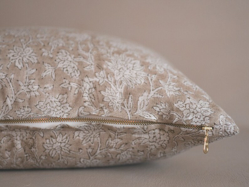 Puede incluir: Una almohada beige con un estampado floral blanco y una cremallera dorada. La almohada est&aacute; hecha de una tela suave y texturizada.