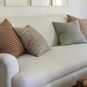 May include: A white linen sofa with a variety of throw pillows. The pillows are brown with white stripes, brown with a floral pattern, and green with a textured pattern.