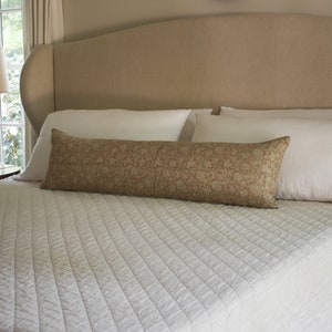 Puede incluir: Una cama blanca con una cabecera beige y un edred&oacute;n acolchado blanco. Una almohada de apoyo de color marr&oacute;n con estampado est&aacute; sobre la cama.