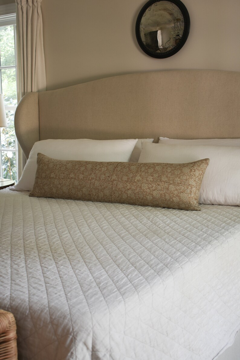 Puede incluir: Una cama blanca con una cabecera beige y un edred&oacute;n acolchado blanco. Una almohada de apoyo de color marr&oacute;n con estampado est&aacute; sobre la cama.