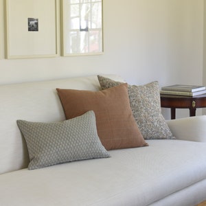 May include: A white couch with a variety of throw pillows in different patterns and colors. The pillows are a light blue with a small pattern, a brown with a subtle texture, and a floral pattern in shades of brown and white. The couch is in front of a white wall with two framed pictures.
