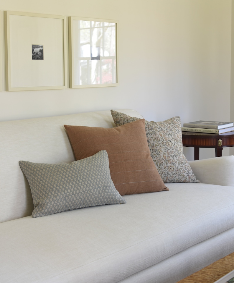 May include: A white couch with a variety of throw pillows in different patterns and colors. The pillows are a light blue with a small pattern, a brown with a subtle texture, and a floral pattern in shades of brown and white. The couch is in front of a white wall with two framed pictures.