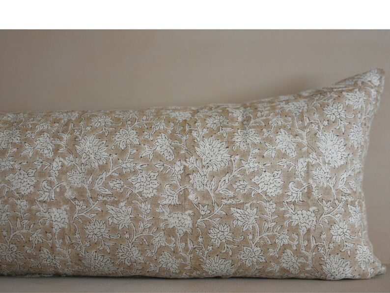 Puede incluir: Una almohada larga y rectangular con una funda de tela de lino beige. La almohada tiene un patr&oacute;n floral blanco con un dise&ntilde;o repetitivo de peque&ntilde;as flores y hojas.