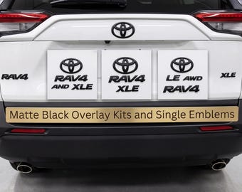 May include: The rear of a white Toyota RAV4 vehicle featuring matte black emblem overlays. Three white square signs display the Toyota logo with text reading RAV4 AND XLE, RAV4 XLE, and LE AWD RAV4. A gold sign reads Matte Black Overlay Kits and Single Emblems.
