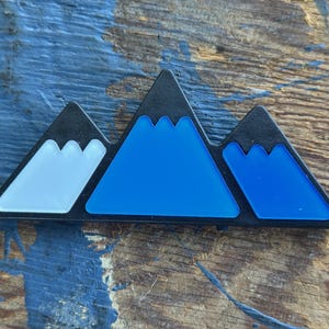 May include: A black mountain range design with three peaks. The peaks are filled with white, blue, and dark blue colors. The design is set against a wooden background.