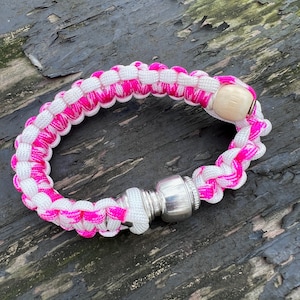 May include: A pink and white paracord bracelet with a wooden bead and a metal attachment.