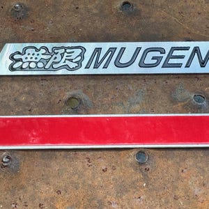 May include: Two rectangular metal car emblems. The top emblem is silver with the word "MUGEN" in black and Japanese characters. The bottom emblem is red with a silver border. Both are on a rusty metal surface.