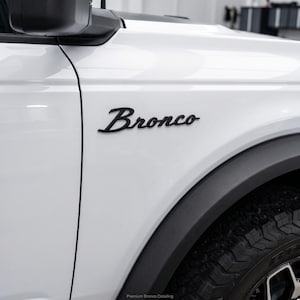 May include: A white vehicle side panel featuring a matte black Bronco emblem in a cursive script font. The panel includes a black plastic wheel arch trim and a portion of a black tire. The text Premium Bronco Detailing is visible at the bottom.