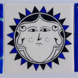 Può includere: Tre piastrelle quadrate in ceramica disposte fianco a fianco. La piastrella a sinistra presenta uno sfondo blu con motivi floreali vorticosi viola e turchesi. La piastrella centrale mostra un volto di sole in bianco e nero con raggi blu. La piastrella a destra mostra un design a forma di cuore blu, giallo e bianco con intricati ghirigori.