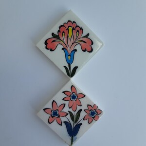 Op de afbeelding: Twee ruitvormige keramische tegels met een witte achtergrond. De bovenste tegel toont een enkele grote roze bloem met gele en blauwe accenten. De onderste tegel toont drie kleinere roze bloemen met blauwe harten en groene stengels.