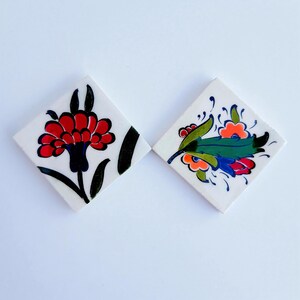 Op de afbeelding: Twee vierkante keramische tegels met een witte achtergrond. De linker tegel heeft een grote rode bloem met donkerblauwe en zwarte accenten. De rechter tegel toont een kleurrijk bloemstuk met groene bladeren, oranje en rode bloemblaadjes en blauwe details.