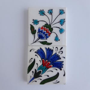 Op de afbeelding: Twee vierkante keramische tegels met een witte achtergrond. De bovenste tegel heeft een centraal bloemmotief met blauw, rood en groen geschilderde bloemblaadjes en stengels. De onderste tegel toont een grote blauwe en rode bloem met groene bladeren.