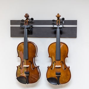 May include: Two brown violins hanging on a black wall mount. The violins have black strings and are in good condition.