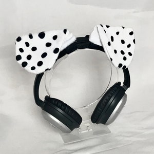 May include: Black and silver over-ear headphones featuring a pair of white plush animal ears with black polka dots attached to the top headband. The headphones have black padded ear cups and a metallic silver outer casing on each side.