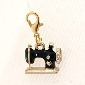 May include: A gold-colored charm featuring a miniature black sewing machine with silver accents. The charm has a gold-colored clasp for easy attachment. The sewing machine has a heart-shaped design and is embellished with small, clear crystals.
