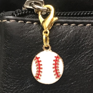 May include: A baseball charm with a gold-colored clasp and a white baseball design with red stitching. The charm is attached to a black leather bag with a silver zipper.