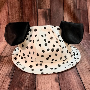 May include: A white bucket hat with black polka dots and two black ears attached. The hat is designed to resemble a Dalmatian dog. The hat is made of a lightweight fabric and is suitable for costume parties or themed events.