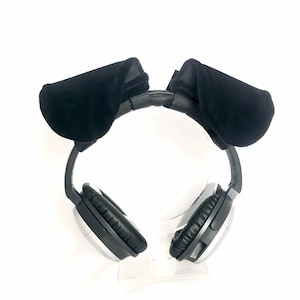 May include: Black headphones with silver accents and black ear pads. The headphones are adorned with plush, black dog ears attached to the headband. The headphones are displayed on a clear stand against a white background.