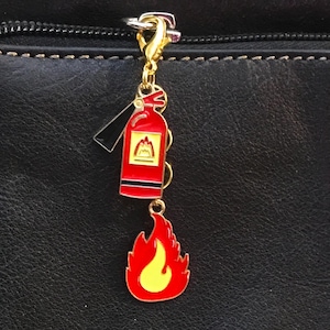 May include: A gold-colored charm with a red fire extinguisher and a red and yellow flame. The fire extinguisher charm has a black handle and a yellow emblem. The charm is attached to a gold-colored clasp.