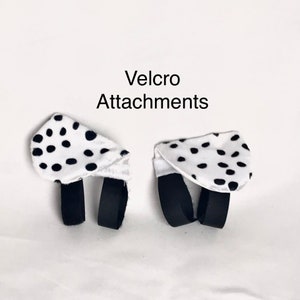 K&ouml;nnte beinhalten: Zwei wei&szlig;e Stoffteile mit schwarzen Punkten, die jeweils zwei schwarze Stoffschlaufen an der Unterseite aufweisen. Der Text 'Velcro Attachments' ist in Schwarz &uuml;ber den Gegenst&auml;nden gedruckt, die zur Befestigung an anderen Objekten dienen.