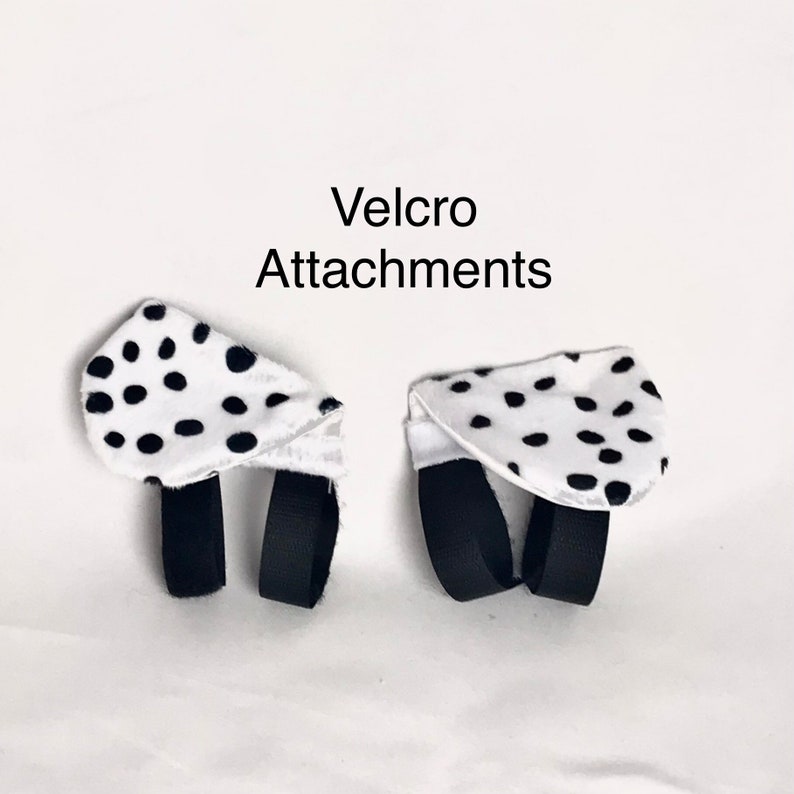 K&ouml;nnte beinhalten: Zwei wei&szlig;e Stoffteile mit schwarzen Punkten, die jeweils zwei schwarze Stoffschlaufen an der Unterseite aufweisen. Der Text 'Velcro Attachments' ist in Schwarz &uuml;ber den Gegenst&auml;nden gedruckt, die zur Befestigung an anderen Objekten dienen.