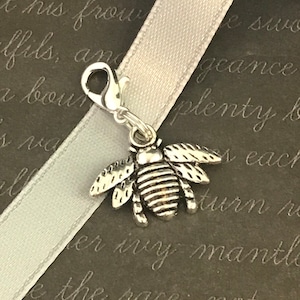 May include: A silver-colored bee charm with detailed wings and striped body, attached to a clasp. The charm is displayed on a white ribbon against a dark background with script text.