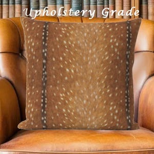 May include: Square decorative pillow with a brown and white spotted pattern, resembling animal hide. The pillow is labeled "Upholstery Grade" and is placed on a leather chair, suggesting a home decor item.