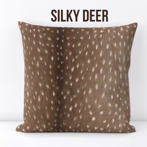 Może przedstawiać: Kwadratowa poduszka z brązowym wzorem i białymi plamkami, przypominająca sierść jelenia. Tekst "SILKY DEER" jest wydrukowany ciemnobrązowym kolorem u g&oacute;ry. Jest to element wystroju domu.