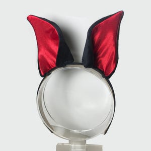 May include: A black headband with two upright ears. The ears are black with red satin interiors. The headband is made of clear plastic. The ears are likely for a costume or themed event.